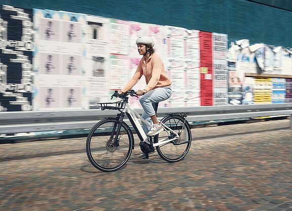 Elektrorad gebraucht Eine Frau fähet auf einem weißen Elektrorad vor einer Wand mit vielen Plakaten.